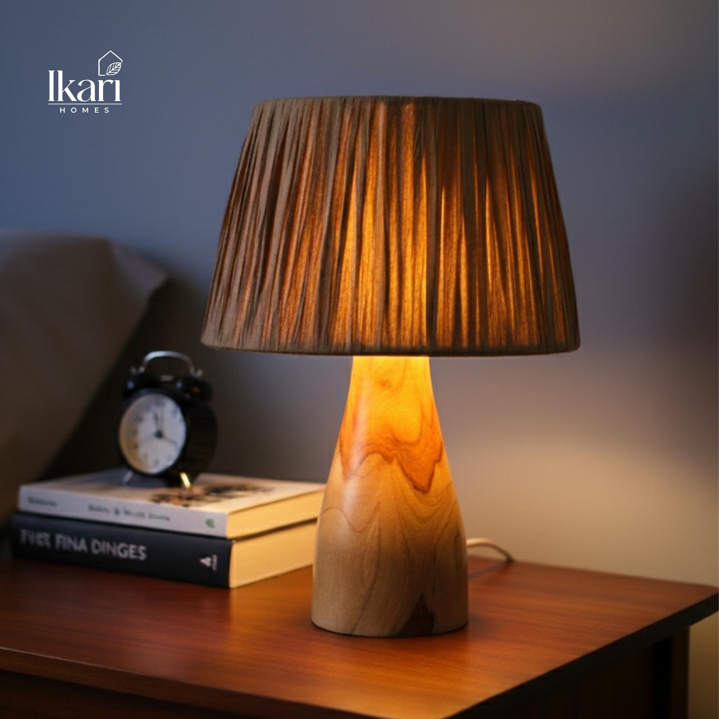 Wooden table lamp with pleated brown shade on a wooden surface, next to books and an alarm clock, with 'Ikari Homes' logo.