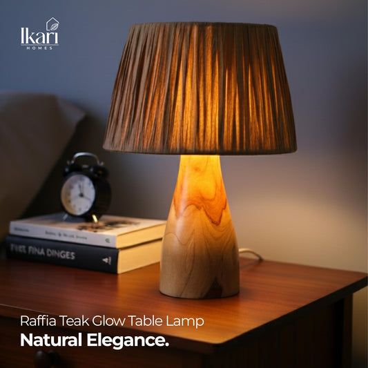 Raffia Teak Glow Table Lamp on a wooden surface with books and an alarm clock in the background, featuring the brand 'Ikari Homes'.
