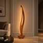 A modern sculptural floor lamp with a wavy wooden frame and warm LED lighting, placed in a stylishly decorated living room next to a sofa.