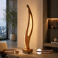 A modern wave-shaped floor lamp with a warm glowing LED light inside a sculptural wooden frame, standing on a square wooden base in a stylishly decorated room. Text overlays highlight its 'Anti-Slip Base' and 'Safety Shockproof Braided Wire' features.