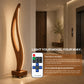 A tall sculptural wooden floor lamp with a curved illuminated wood body stands in a modern hallway. A smartphone remote control is shown beside it, with marketing text reading 'LIGHT YOUR MOOD, YOUR WAY.